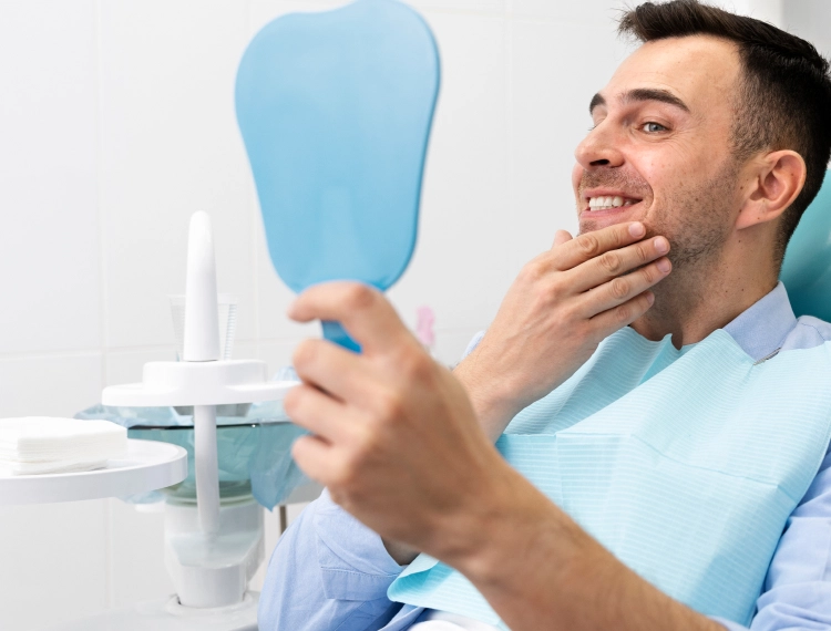 Smiling man sitting in a dental chair holding a blue hand mirror and touching his chin.